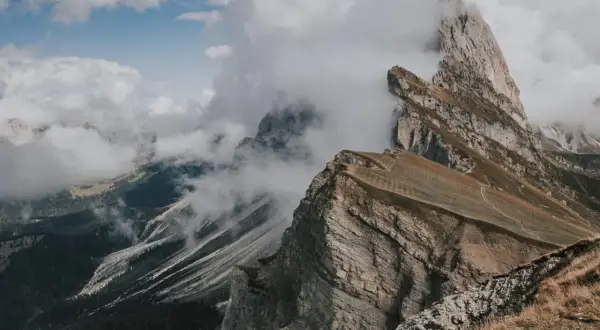 Molnen sveper in över dramatiska bergstoppar i Dolomiterna, Italien. En vandringsled slingrar sig uppför en gräsklädd sluttning.