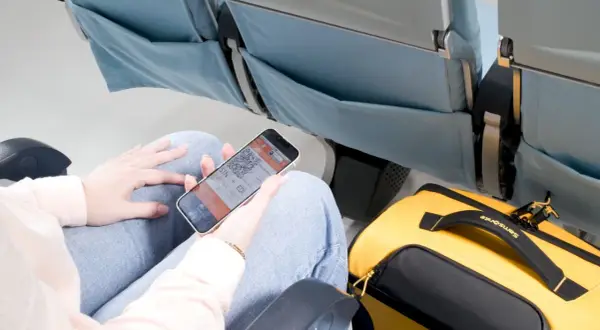 Person visar boardingkort på mobilen i ett flygplanssäte. Gul Samsonite-väska bredvid. Smidigt med digitala biljetter för ditt ving flygbolag.