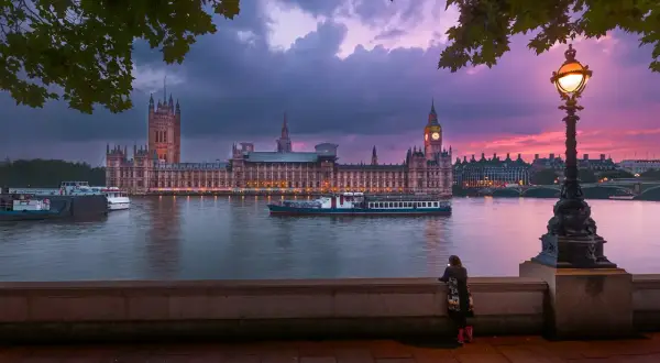 En kvinna ser på Houses of Parliament vid solnedgången. En båt glider förbi på Themsen, en ikonisk vy över London stadsdelar.