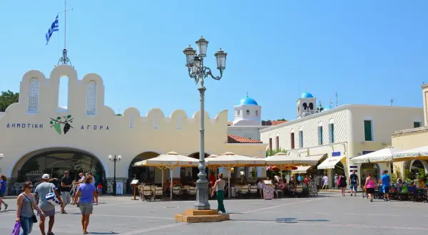 Solig dag på **Kos stad**s torg. Folk sitter vid caféer under parasoller framför en gul byggnad med grekisk flagga. Blå kupoler syns i bakgrunden.
