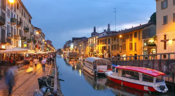 Kvällsliv vid Navigli i Milano. Folk promenerar längs kanalen, båtar ligger vid kaj och restauranger lyser upp.