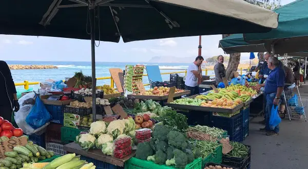 Frukt- och grönsaksstånd vid marknad i Chania. Färska varor som majs, broccoli och tomater säljs med havet i bakgrunden.