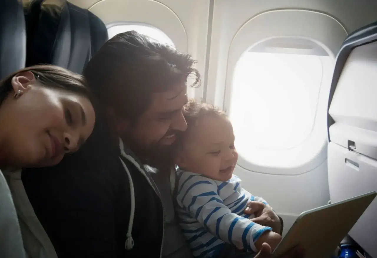 En familj njuter av att flyga med barn. Pappan håller sitt leende barn som tittar på en surfplatta, medan mamman vilar sitt huvud mot hans axel.