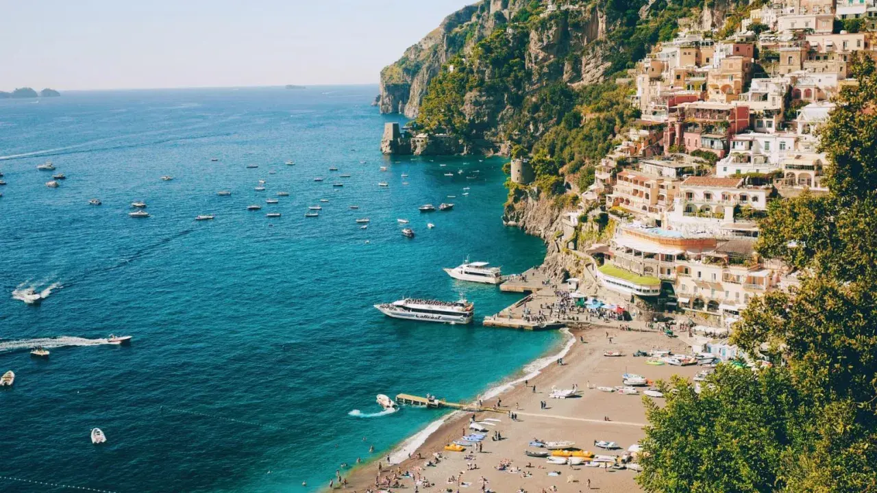 Färgstark by klättrar uppför klipporna vid sorrentohalvön, med båtar på det turkosa havet och en strand full av folk.