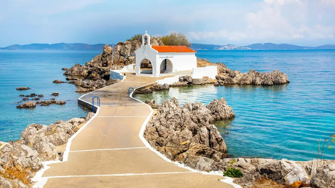 En vit kyrka med orange tak på en klippig ö, nås via en gångväg. Vackert vid Skiathos, Grekland.