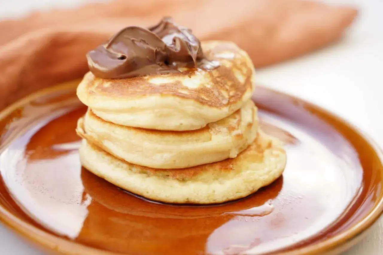 En stapel fluffiga amerikanska pannkakor toppas med chokladkräm på en brun tallrik.