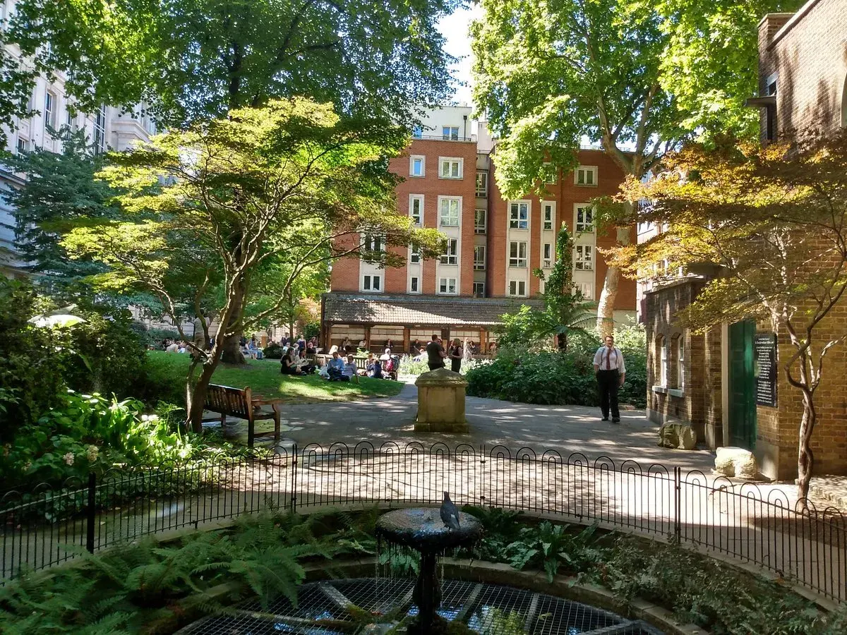 En grönskande park i London med människor som kopplar av på gräsmattan. En fågel sitter på en fontän i förgrunden.