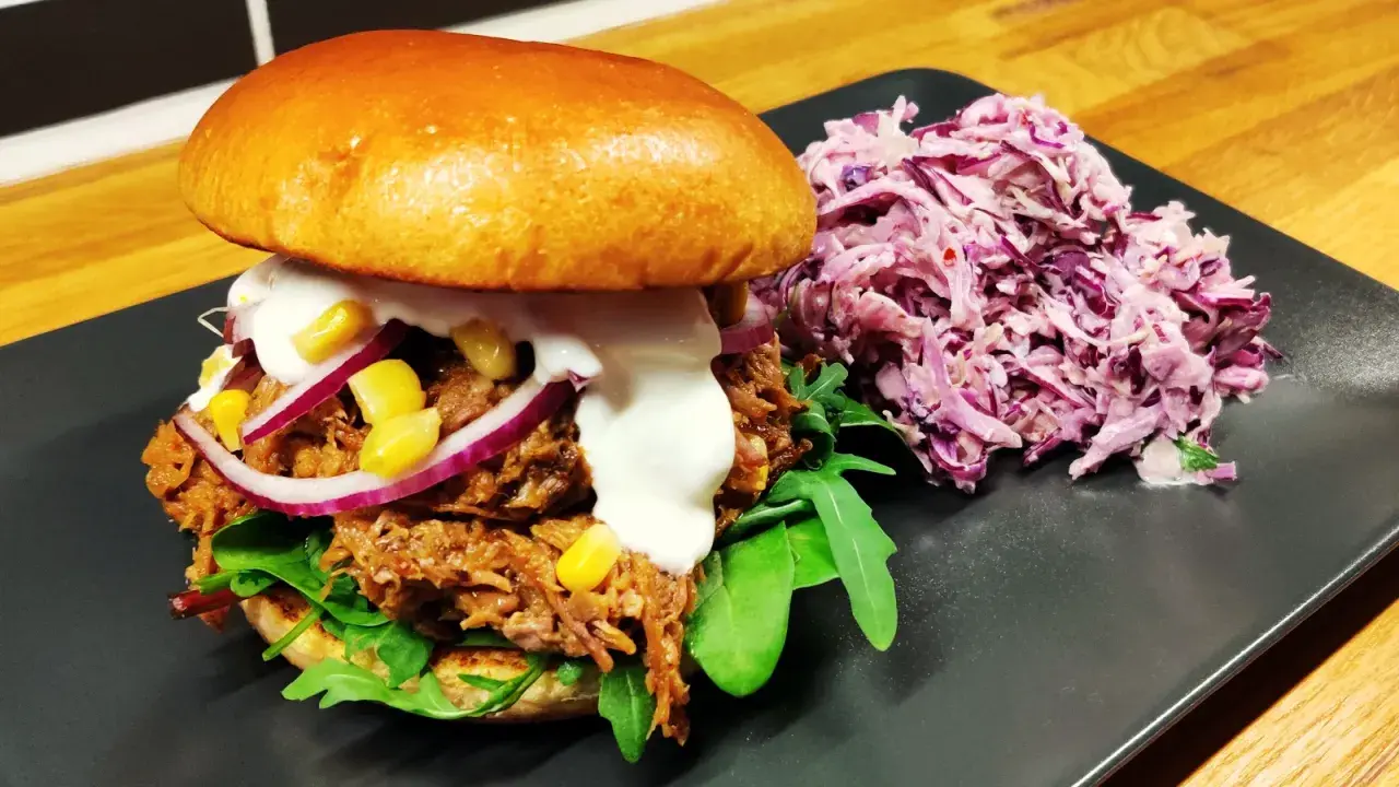En saftig pulled pork-burgare med majs, rödlök och krämig sås, serverad med en lila kålsallad.