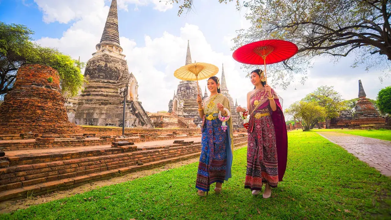 Två kvinnor i traditionella thailändska kläder med parasoller promenerar vid gamla tempelruiner, en scen som påminner om den kultur som talas i Bangkok.