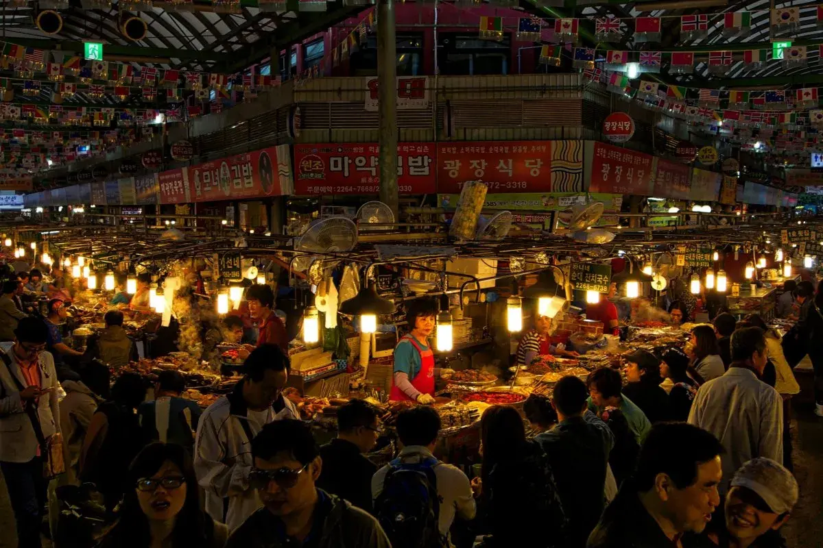 Livligt folkliv på Gwangjang Market, med matstånd som säljer koreansk mat under blinkande lampor och flaggor.