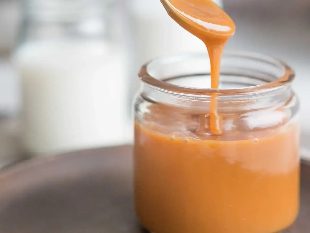 A spoon pours smooth, glossy caramel sauce into a glass jar.