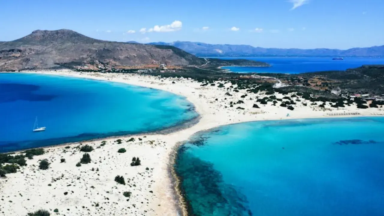 En vit sandstrand med turkost vatten och en segelbåt vid **Platanias, Kreta**. Berg i bakgrunden.