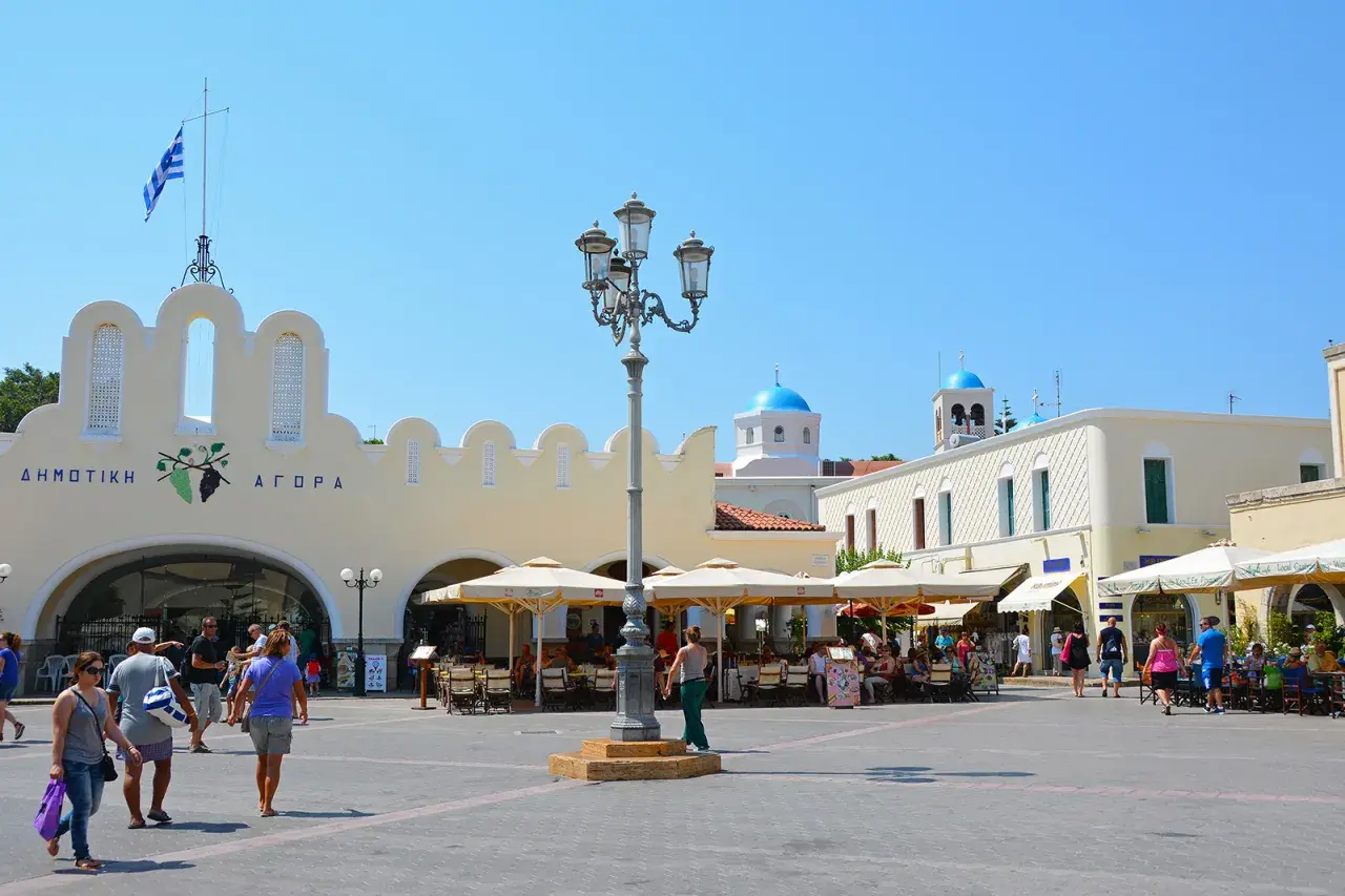 Solig dag på **Kos stad**s torg. Folk sitter vid caféer under parasoller framför en gul byggnad med grekisk flagga. Blå kupoler syns i bakgrunden.
