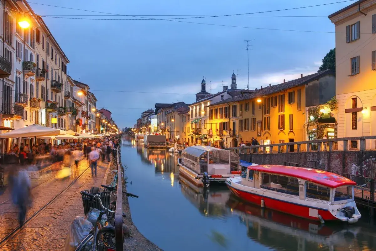 Kvällsliv vid Navigli i Milano. Folk promenerar längs kanalen, båtar ligger vid kaj och restauranger lyser upp.