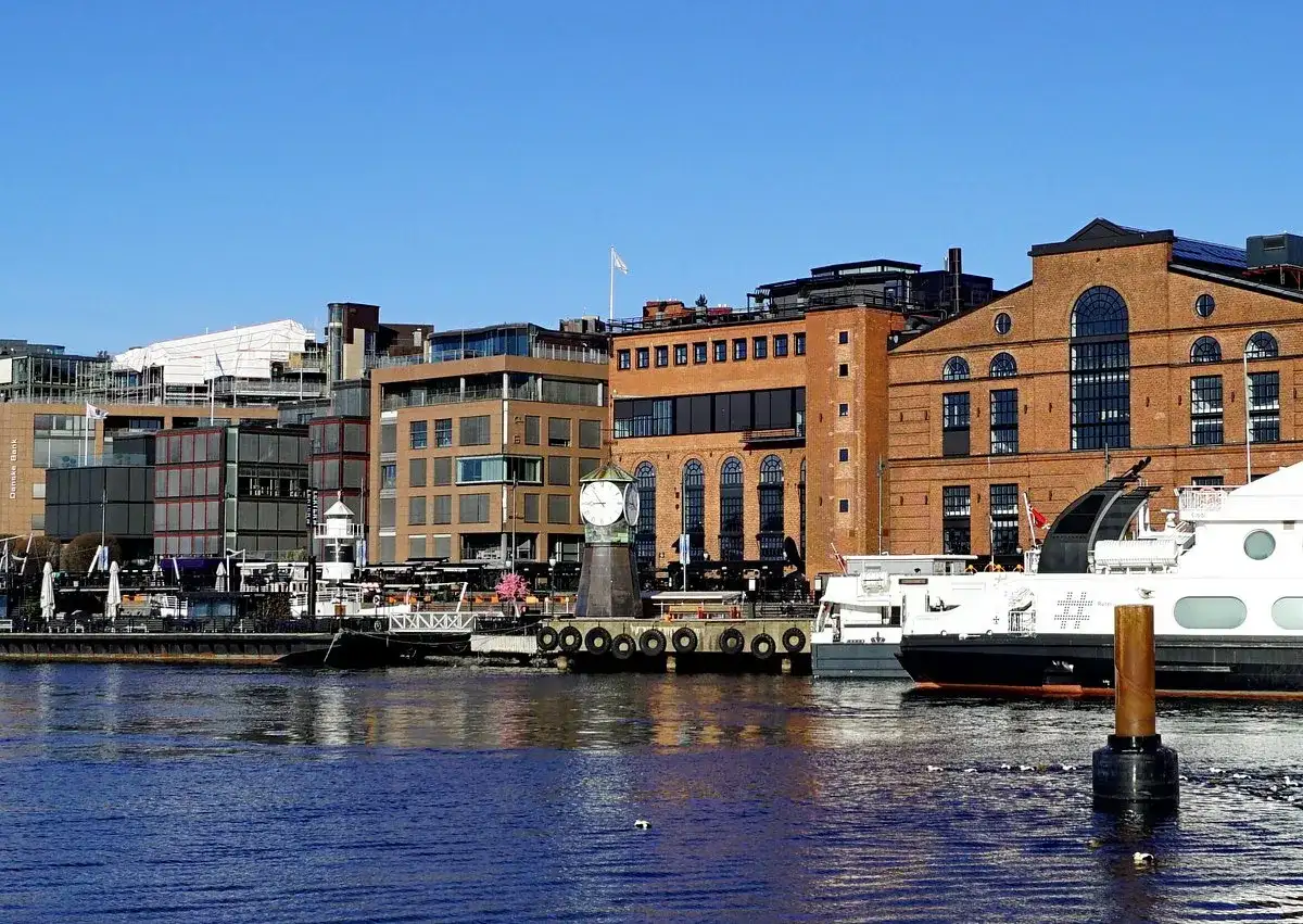 En del av Oslo med moderna byggnader, en gammal tegelbyggnad med ett stort klocktorn vid vattnet och båtar.