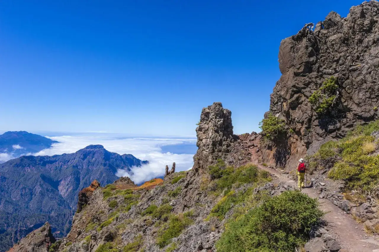 En person vandrar p&aring; Gran Canaria, med dramatiska klippor och molnhav under en klarbl&aring; himmel.