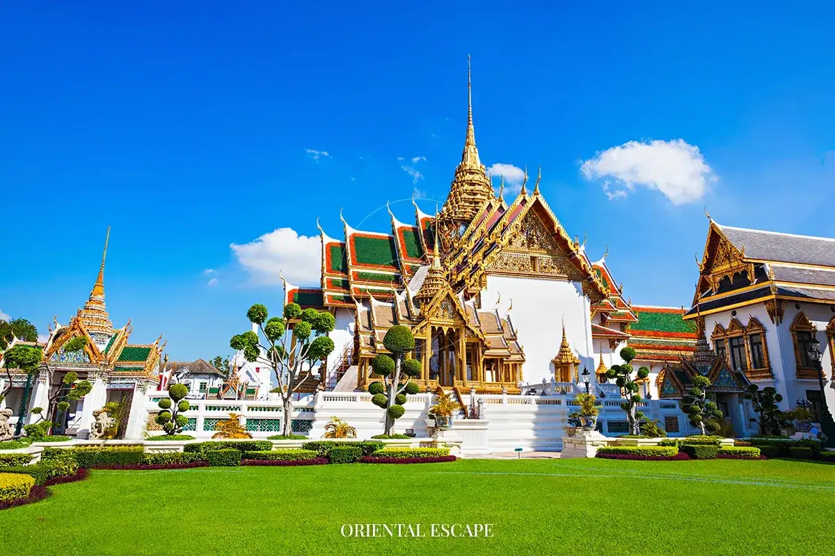 Ett magnifikt tempel i Bangkok med gyllene tak och gr&ouml;na spiror, omgivet av en v&auml;lsk&ouml;tt gr&auml;smatta och tr&auml;d.