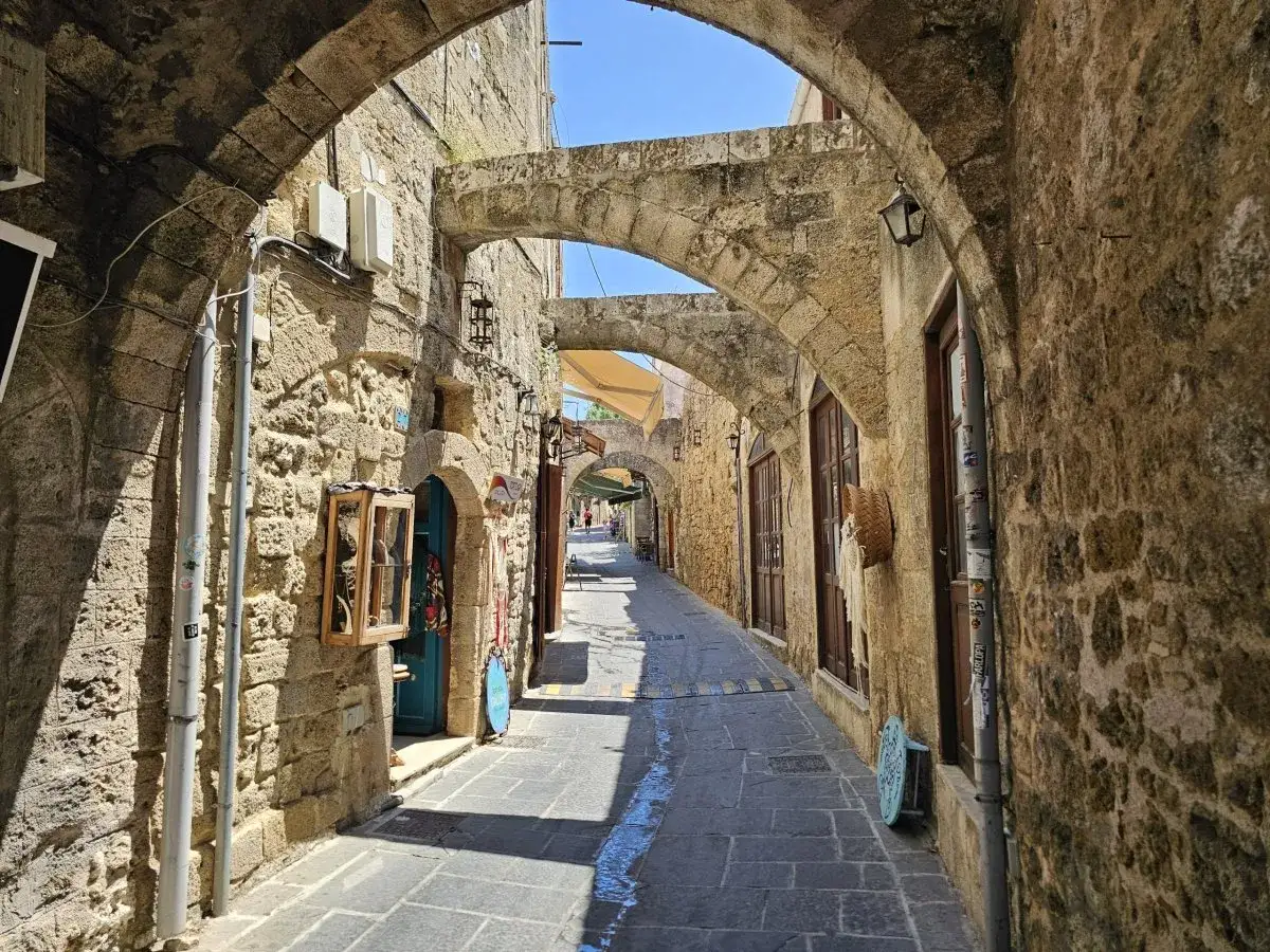 En smal, stenbelagd gr&auml;nd i Rhodos stad, kantad av gamla stenhus med valv och tr&auml;f&ouml;nster. Solen kastar l&aring;nga skuggor.