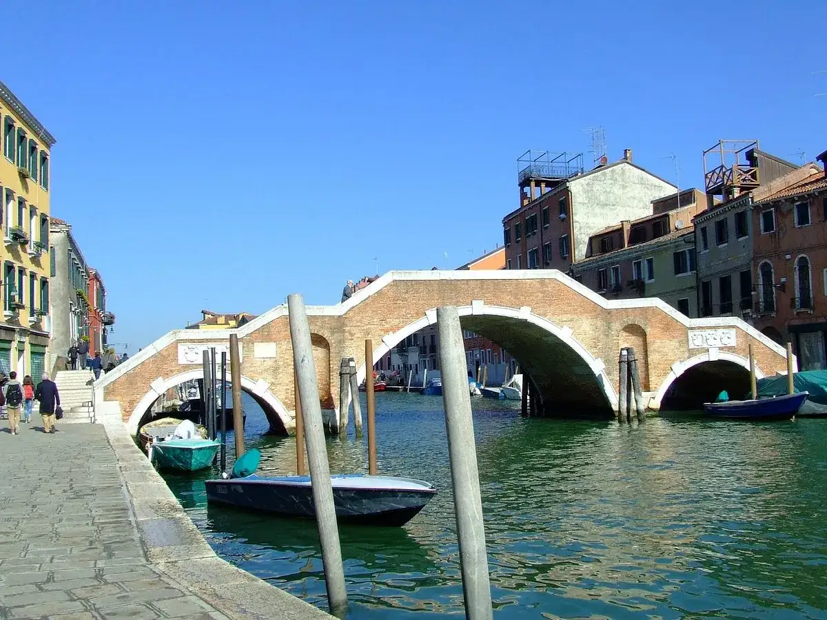 En vacker vy av en del av Venedig med en tegelbro &ouml;ver en kanal, d&auml;r sm&aring; b&aring;tar guppar i det gr&ouml;na vattnet.