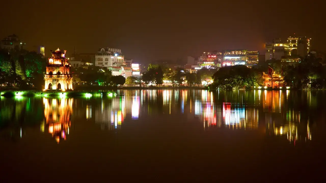Nattvy &ouml;ver Hoan Kiem-sj&ouml;n i Hanoi, huvudstad i Vietnam. Upplysta byggnader och ett torn speglas i vattnet.
