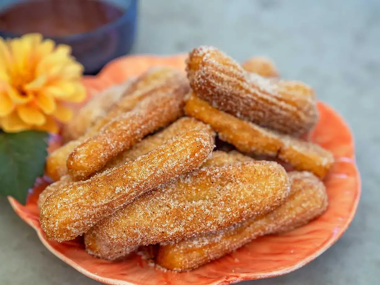 En h&ouml;g med socker- och kaneldr&auml;nkta churros p&aring; en orange tallrik. Perfekt f&ouml;r ett enkelt churros recept.