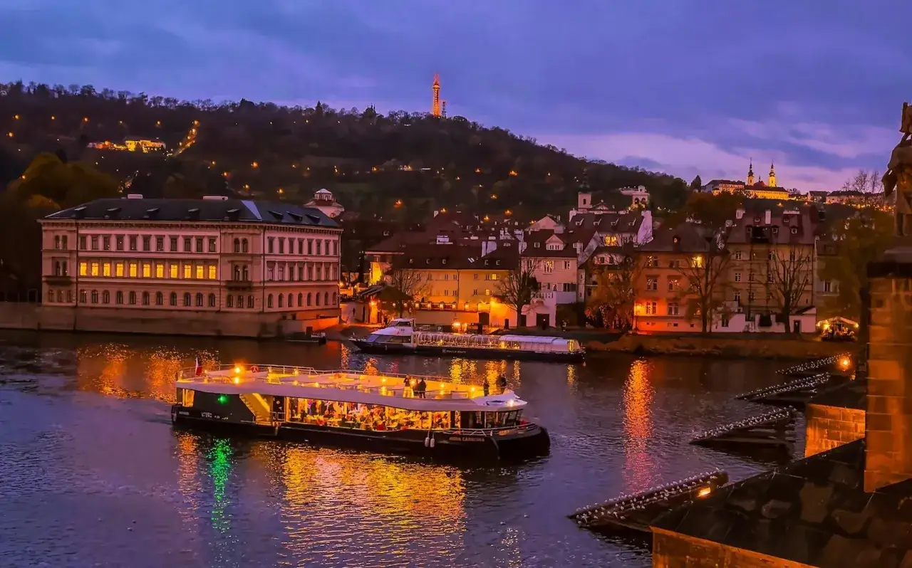 Kv&auml;llsb&aring;t p&aring; Moldau i Prag, med upplysta byggnader och kullar i bakgrunden. Perfekt f&ouml;r att uppt&auml;cka Prags b&auml;sta barer.