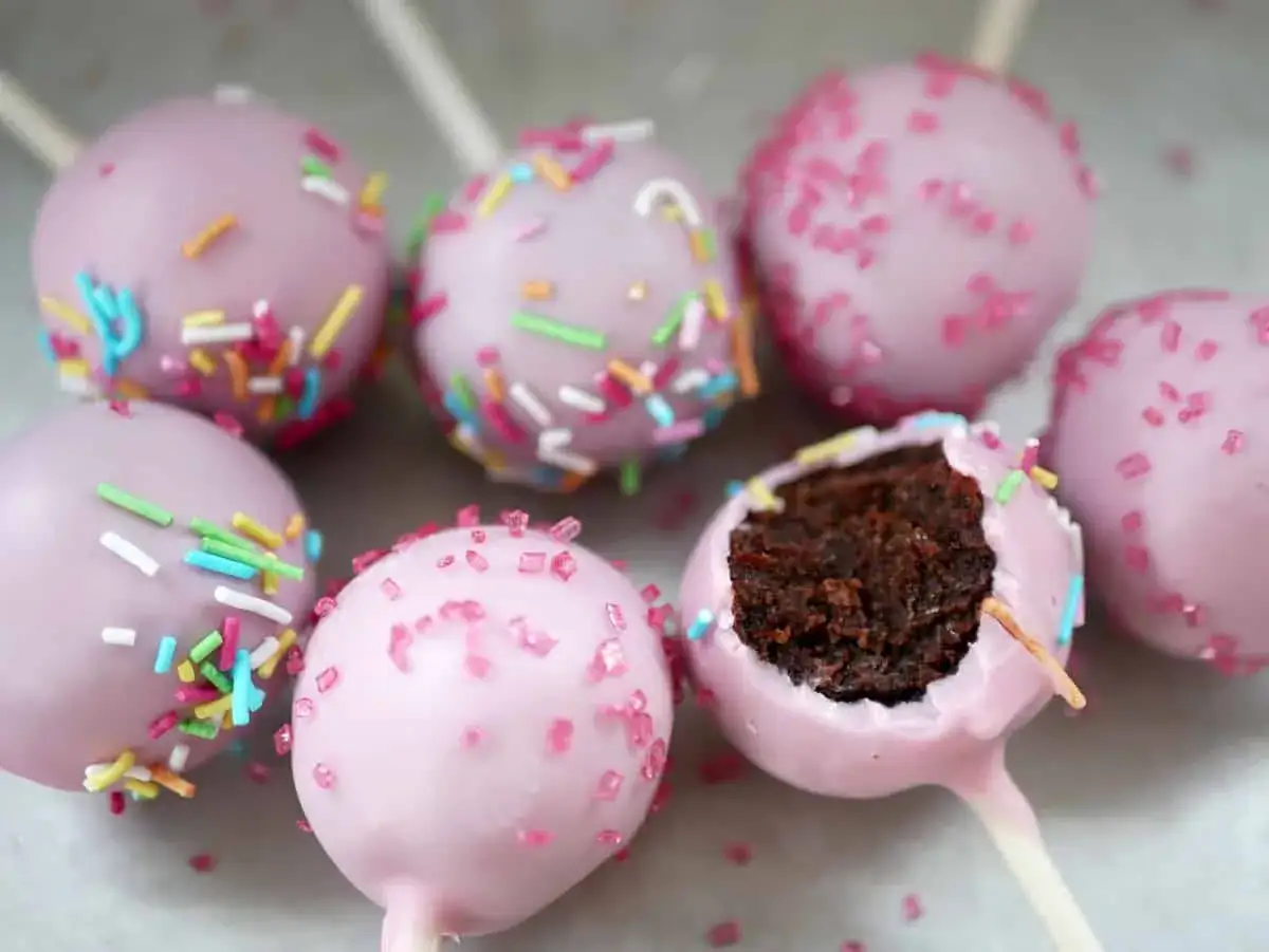 Sju rosa cake pops med str&ouml;ssel. En &auml;r upp&auml;ten och visar det m&ouml;rka chokladinneh&aring;llet.