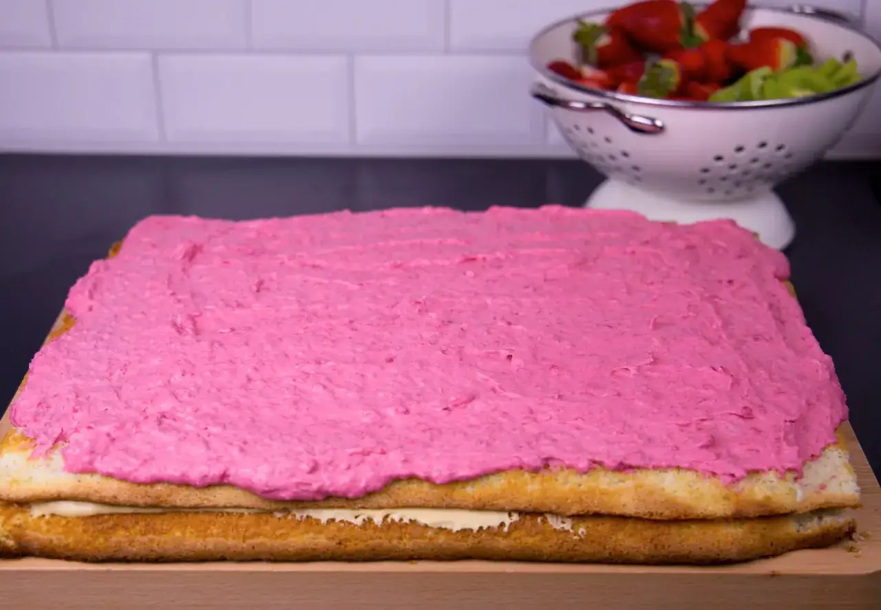 En rosa frosting t&auml;cker en t&aring;rtbotten i l&aring;ngpanna, redo f&ouml;r dekoration. Jordgubbar och kiwi syns i bakgrunden.