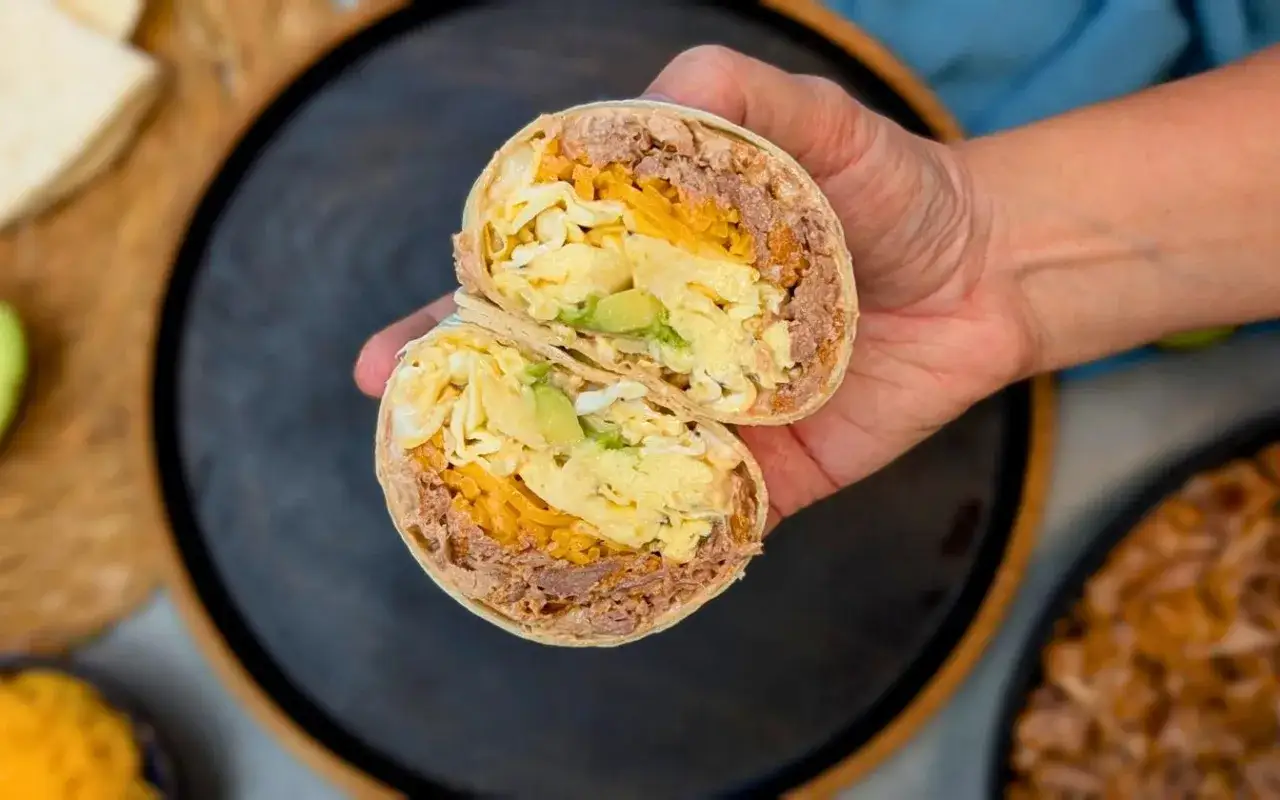 En hand h&aring;ller en delad frukost burrito med &auml;gg, avokado, ost och k&ouml;tt. Perfekt f&ouml;r en m&auml;ttande start p&aring; dagen!