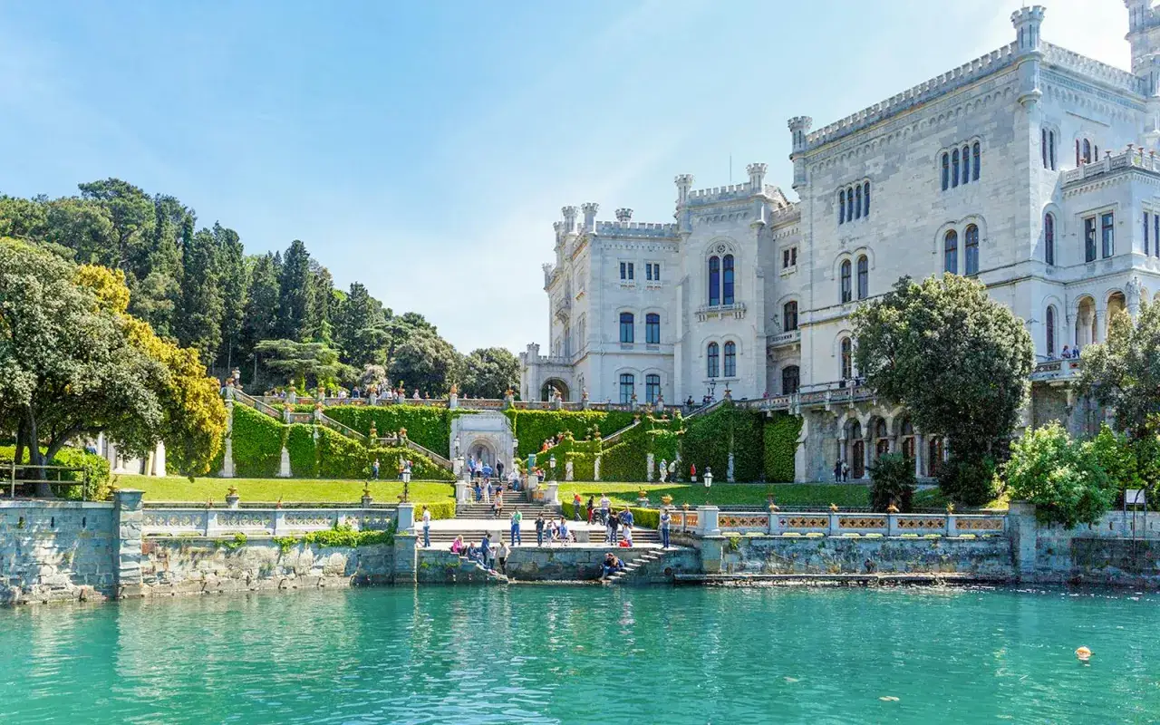 Vackert slott vid havet i Trieste, Italien. Gr&ouml;nskande tr&auml;dg&aring;rdar och turkost vatten.