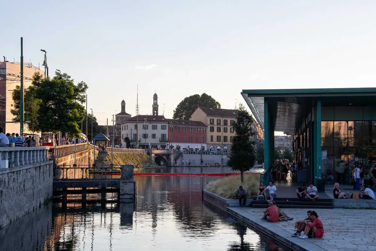 M&auml;nniskor njuter av kv&auml;llssolen vid Navigli i Milano. Vatten reflekterar f&auml;rgglada byggnader och en modern gr&ouml;n byggnad.