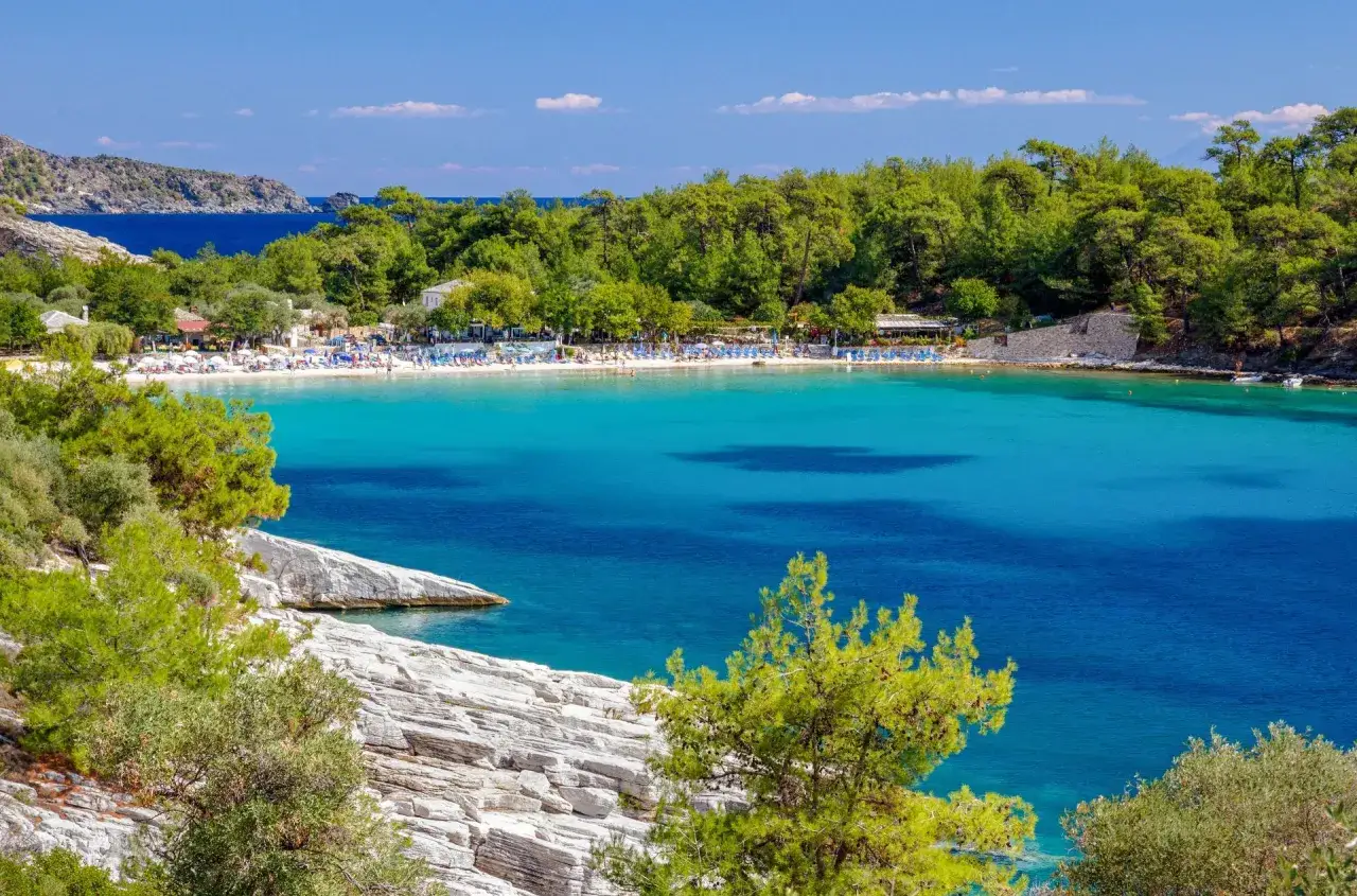 En vacker vy &ouml;ver en turkos vik med en strand full av solstolar och parasoller p&aring; Thassos, Grekland. Gr&ouml;nskande tr&auml;d och klippor ramar in scenen.