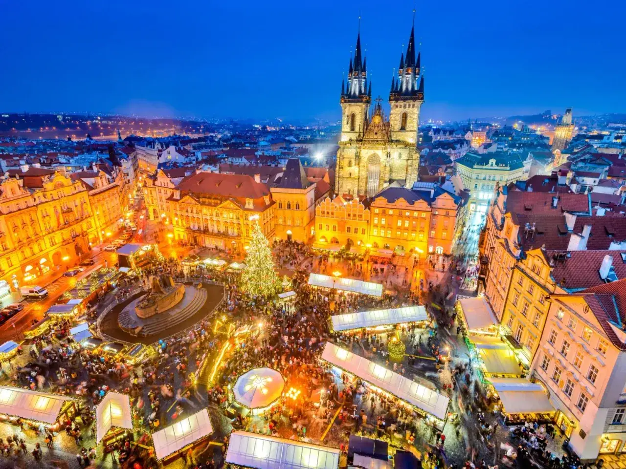 En livlig prag julmarknad med en stor julgran och m&aring;nga bes&ouml;kare. Gamla byggnader och kyrktorn lyser upp kv&auml;llen.