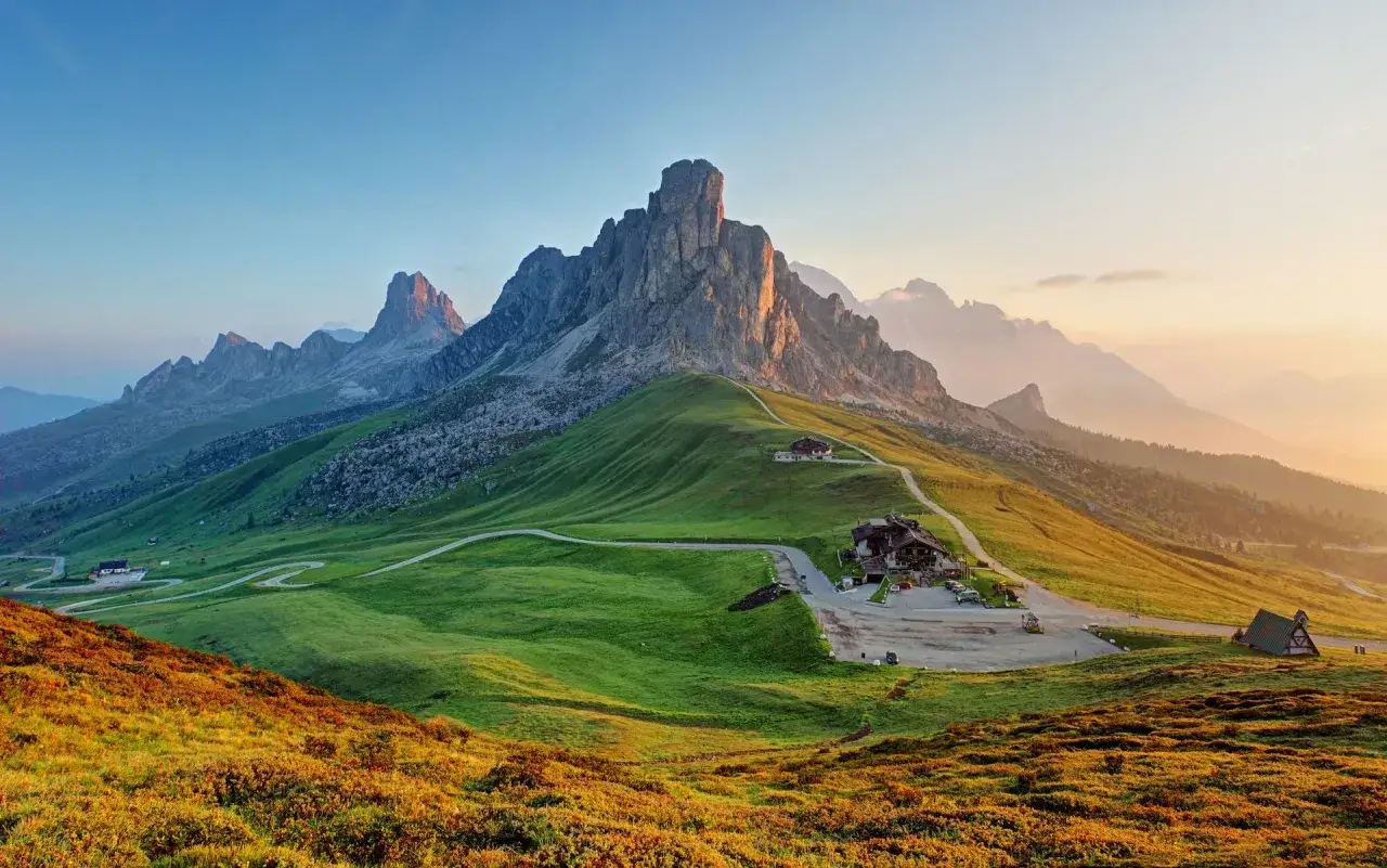 Solnedg&aring;ng &ouml;ver Dolomiterna i Italien. Gr&ouml;nskande kullar med slingrande v&auml;gar och bergstoppar som badar i gyllene ljus.