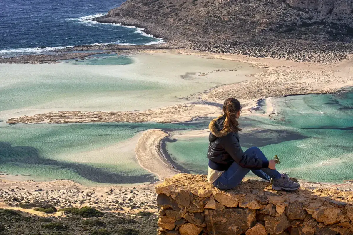 En kvinna sitter p&aring; en klippa och njuter av utsikten &ouml;ver turkost vatten och sandbankar. En perfekt vy att se p&aring; Kreta.