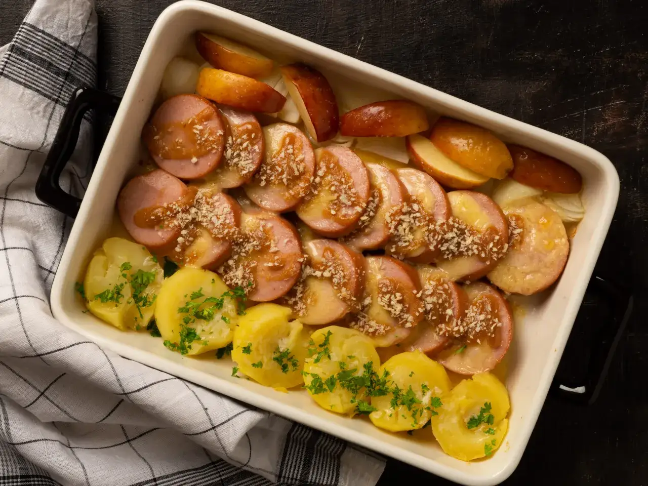 En ugnsform med skivad falukorv, potatis, &auml;pple och l&ouml;k. R&auml;tten &auml;r toppad med en s&aring;s och str&ouml;br&ouml;d.