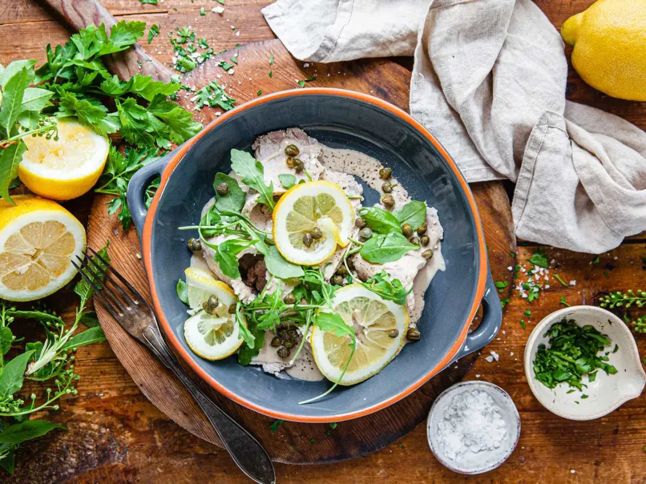 Vitello tonnato i en gr&aring; sk&aring;l, toppad med citronskivor, kapris och ruccola. Serveras med citroner, f&auml;rska &ouml;rter och salt p&aring; ett tr&auml;bord.