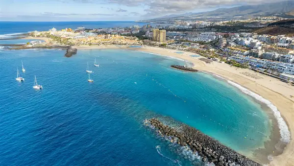 Vacker vy över en strand med turkost vatten och segelbåtar. En stad breder ut sig längs kusten, perfekt för en resa till Teneriffa i november.