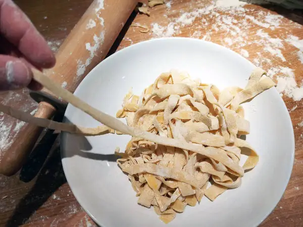 Färska tagliatelle av pastadeg durumvete läggs i en skål. En kavel och mjöl syns i bakgrunden.