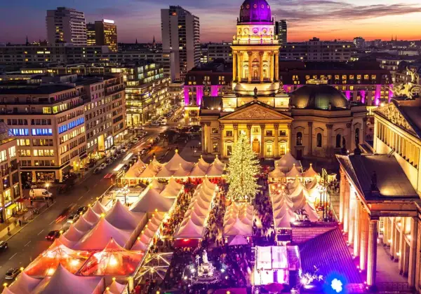 En livlig julmarknad i Berlin med tält, en stor julgran och upplysta byggnader i skymningen.