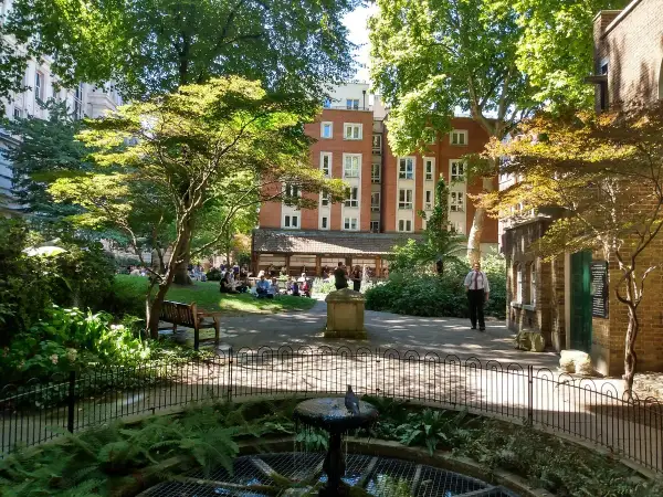 En grönskande park i London med människor som kopplar av på gräsmattan. En fågel sitter på en fontän i förgrunden.