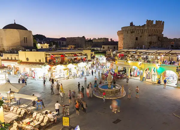 Kvällsliv på Rhodos med folk som njuter av Rhodos shopping och restauranger vid en fontän, med en borg i bakgrunden.