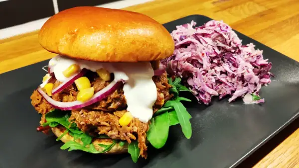En saftig pulled pork-burgare med majs, rödlök och krämig sås, serverad med en lila kålsallad.
