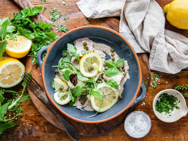 Vitello tonnato i en skål, toppad med citronskivor, kapris och ruccola. Serveras med citroner och färska örter.