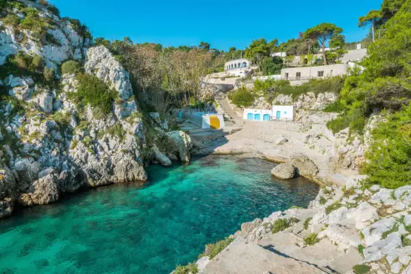 En klippig vik med turkost vatten i Salento, Italien. Vita byggnader med blå dörrar och en gul vägg klättrar uppför sluttningen.