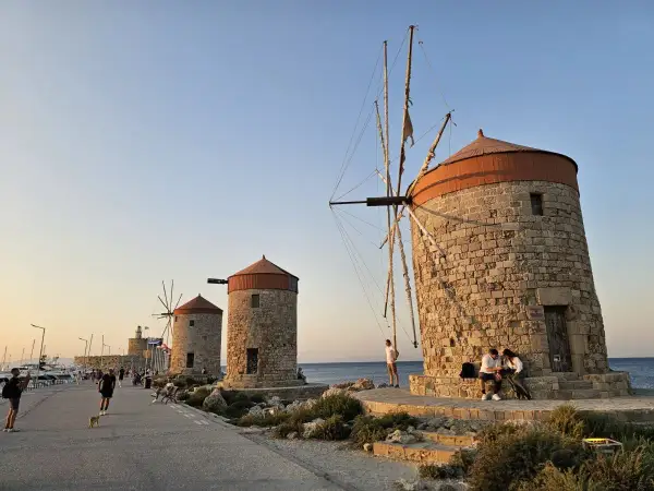 Gamla väderkvarnar längs kajen i Rhodos stad. Folk promenerar och njuter av kvällssolen vid havet.
