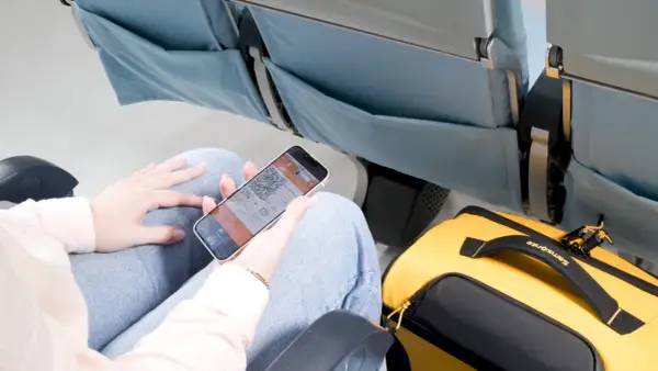 Person visar boardingkort på mobilen i ett flygplanssäte. Gul Samsonite-väska bredvid. Smidigt med digitala biljetter för ditt ving flygbolag.