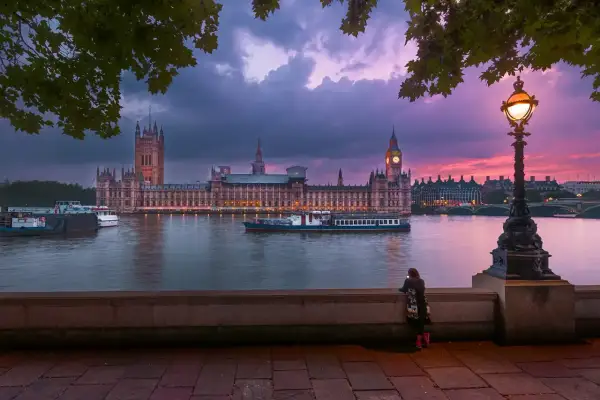 En kvinna ser på Houses of Parliament vid solnedgången. En båt glider förbi på Themsen, en ikonisk vy över London stadsdelar.